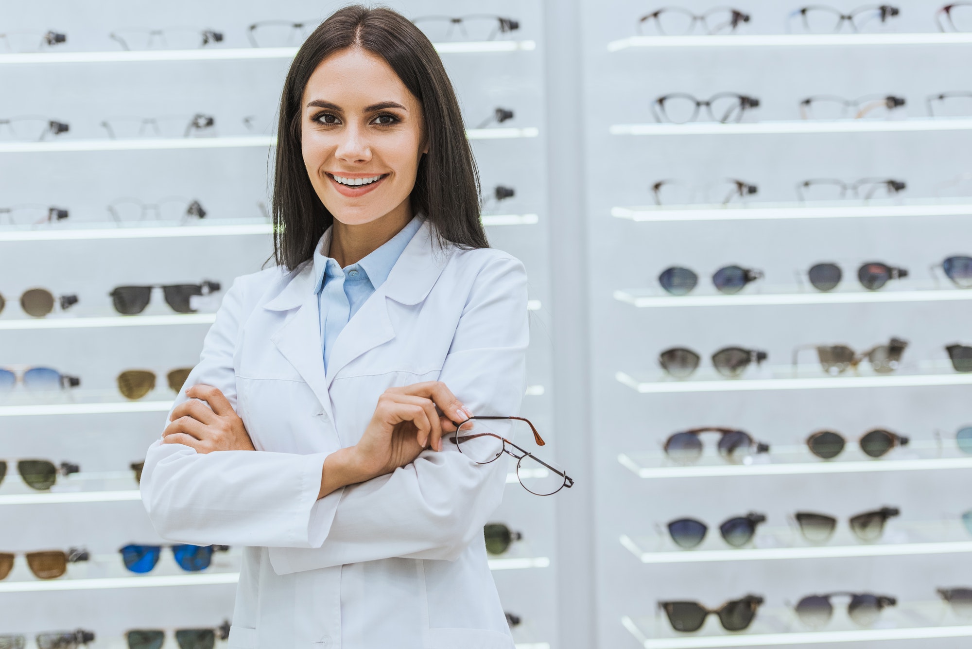 professional smiling optician holding glasses near shelves in optica
